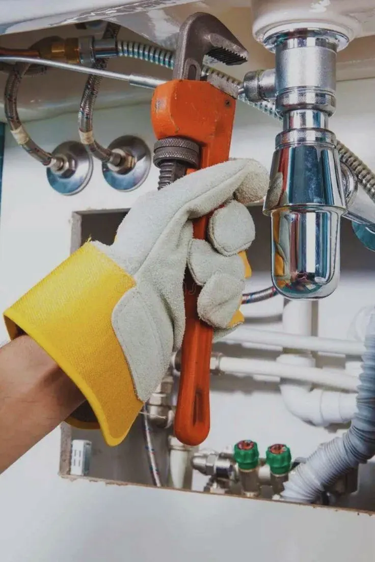 A gloved hand holding a pipe wrench on the pipes under a sink